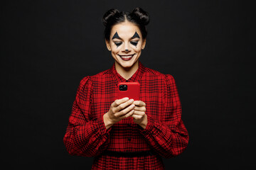 Young woman with Halloween makeup face art mask wear clown costume red dress hold in hand use mobile cell phone isolated on plain solid black background studio portrait. Scary holiday party concept.