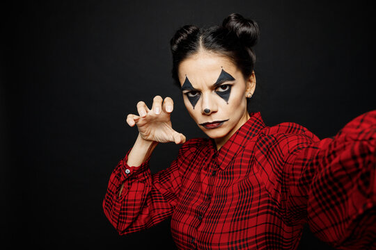 Young woman with Halloween makeup face art mask wears clown costume red dress do creepy selfie shot pov mobile cell phone isolated on plain solid black background studio. Scary holiday party concept.