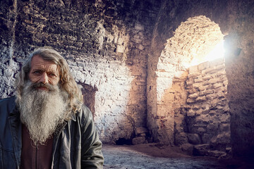 In the subterranean depths of a castle, an old man with a silver beard stands against a stone wall,...