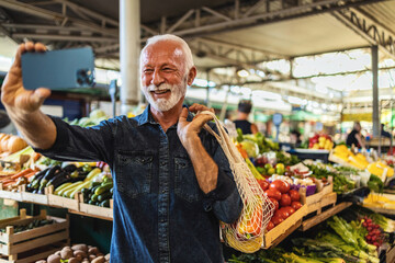 Joyful senior Caucasian man standing at  farmer's market using his mobile phone to take a selfie.