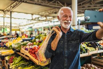 Joyful senior Caucasian man standing at  farmer's market using his mobile phone to take a selfie.