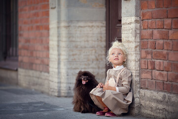 Little girl with blonde hair wearing dress with a dog on the city street 