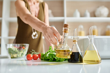 cropped view of woman taking glass bottle with olive oil while preparing salad in kitchen - obrazy, fototapety, plakaty