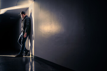 Serious sad man standing in dark corridor. Mystery guy in spotlight. Dramatic light and shadow. Stalker or outcast. Person waiting, hiding, sneaking or eavesdropping. Despair, trauma or sorrow. - obrazy, fototapety, plakaty