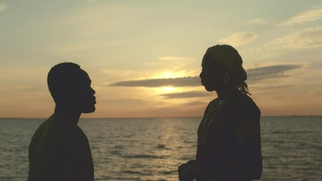 Medium shot of young beautiful African American couple standing by water at golden sunset and having conversation