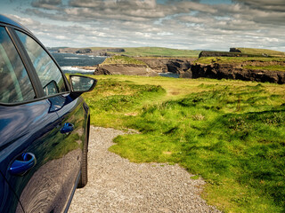 Dark color car parked off road, Stunning nature scene in the background with cliffs and low cloudy sky, sunny day. Kilkee area, Ireland. Travel, tourism and sightseeing concept. Irish landscape. - obrazy, fototapety, plakaty
