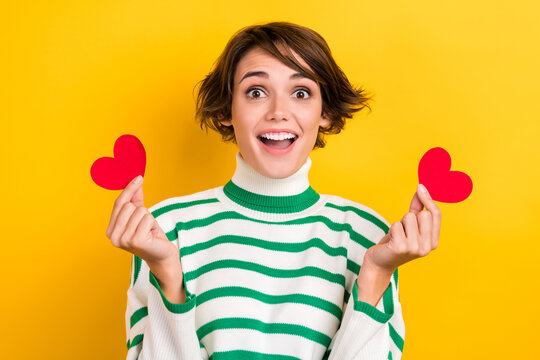Photo of shiny excited lady wear striped pullover holding two small red hearts isolated yellow color background - Powered by Adobe