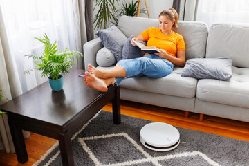 Robotic vacuum cleaner cleaning a room while a woman reading book on the sofa