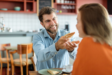 Portrait of smiling european man giving sandwich to his wife, feeding woman while sitting in cafe interior, free space. Nutritious breakfast, snack