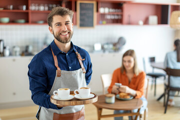 Cheerful male waiter carrying coffee cups on tray and smiling at camera, working in his small business restaurant cafe