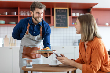 Handsome friendly male waiter serving some pastry to female customer, woman sitting in cafe or coffeeshop