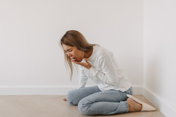 Fototapeta na wymiar Asian Thai woman feeling sick and queasy, cover mouth, has a stomachache cramp in white sweater, sitting on floor in white apartment room in winter.