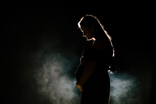 In the low key rim light portrait, the silhouette of a beautiful pregnant woman's belly stands out against a dark black background, creating a striking and evocative image