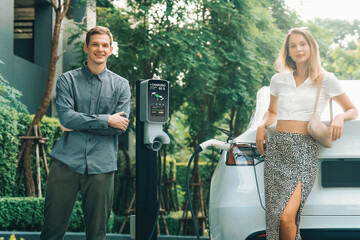 Young couple travel with EV electric car charging in green sustainable city outdoor garden in...