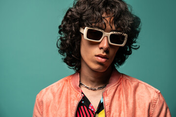 Confident young ethnic man in sunglasses pink jacket standing near turquoise backdrop