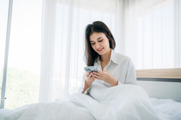 Young Woman Waking Up and play phone in Cozy Bedroom at Home. Beautiful Girl Smiling and Relaxing in Bed after Waking Up.