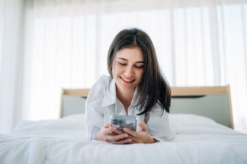 Young Woman Waking Up and play phone in Cozy Bedroom at Home. Beautiful Girl Smiling and Relaxing in Bed after Waking Up.