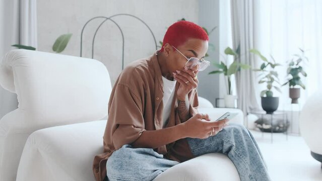 Young shocked joyful African American woman with smartphone in hands feels surge positive emotions reading e-book and learning about happy ending for literary characters sits in white chair in home