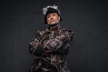 Pirate with a gray beard dressed in a brown vest and hat posing with crossed arms against a...