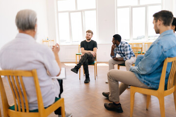 View from back of professional mature male psychologist to depressed young man sharing problem sitting in circle during group therapy session. Concept of mental health, psychotherapy, social issues.