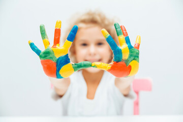 little blonde girl shows her hands painted with multi-colored paint on a white background.