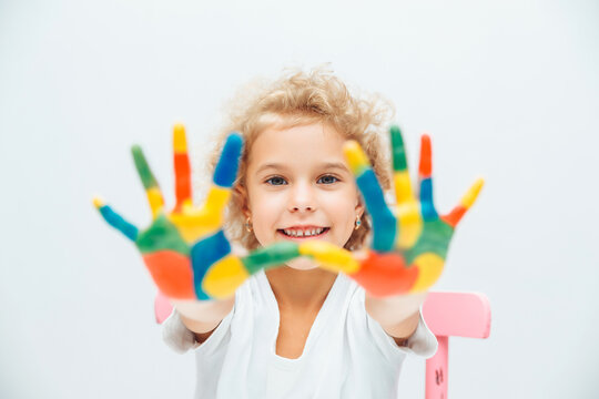 little blonde girl shows her hands painted with multi-colored paint on a white background.