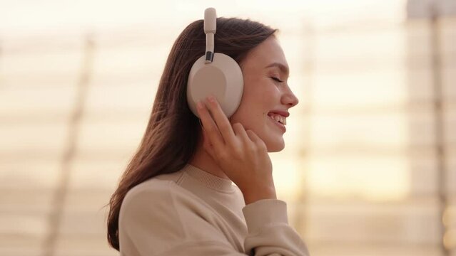 A side view of a teenage girl headbanging to rhythmic music in overhead headphones at sunset, basking in warm light and smiling
