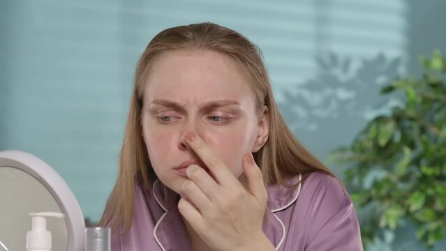 Woman scrutinizes her face in the mirror, forehead, nose. Young woman with problem skin covering her face with her hands crying in frustration.