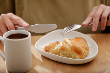 woman's hands cutting a croissant next to a coffee cup on a light wood teapot