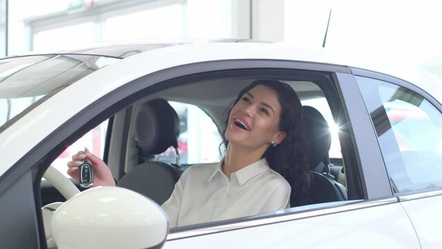 Portrait of a happy woman driving a new luxury electric car in a car showroom. A woman shows emotions of happiness.