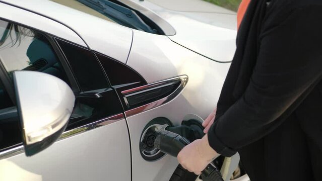 Female hands insert power connector into EV car. Unrecognizable pregnant woman in dress plugging in charging cable to electric vehicle and charges batteries. Connect EV plug