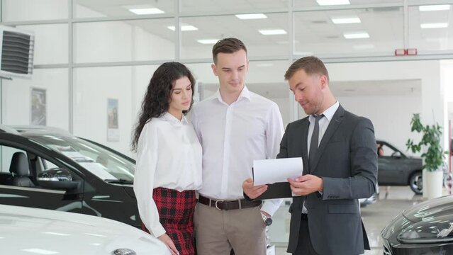 Automobile business, sale of new cars. A happy couple talks to a dealer talking about a car in a car showroom. 