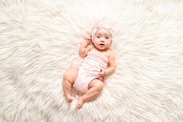 Photograph of an infant lying on a white fluffy blanket. The infant is wearing a pink jumpsuit and a pink headband with a bow