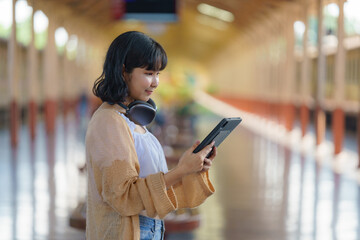 A backpacker is standing on a train station platform, using a tablet to plan her next destination. She has headphones around her neck to listen to music or audiobooks.