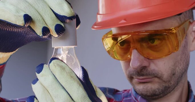 Portrait of a man wearing a safety helmet and goggles on his face, an electrician screws an electric light bulb with a spiral into a lamp socket. Connecting indoor lighting