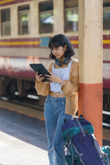 A backpacker is standing on a train station platform, using a tablet to plan her next destination. She has headphones around her neck to listen to music or audiobooks.