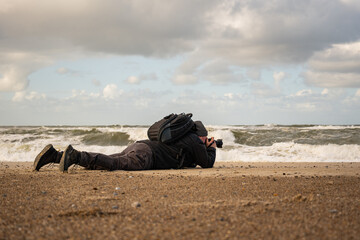 Photographer lies on the beach to photograph the birds above the water and the strong waves. Windy and cloudy day on the Danish North Sea coast near Blavand. Portrait of a wildlife Photographer.