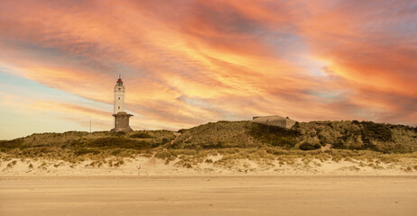 Large lighthouse on the Danish coast in the town of Blavand. Beautiful calm orange blue sky with soft clouds. Dune landscape on the beach in Denmark. Autumn travel destination to relax. Panorama