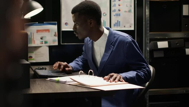 Private investigator seen in dimly lit office meticulously analyzing papers and evidence gathered during research. With laptop open, policeman cross-references information and does background check.