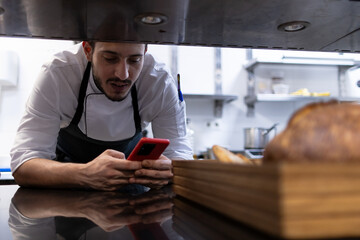 An arab chef is leaning against the kitchen while using his mobile phone.