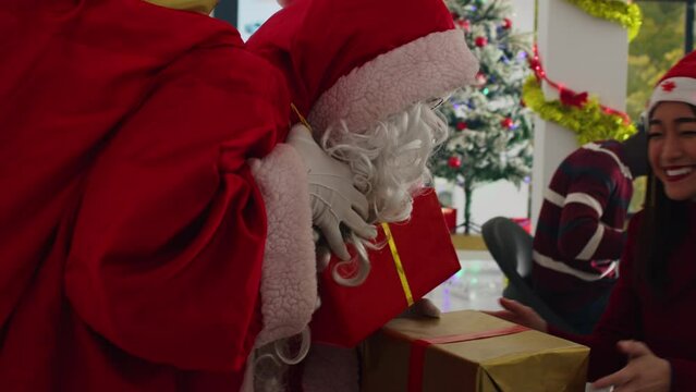Close up shot of manager dressed as Santa Claus spreading holiday joy in festive adorn office, offering presents to employees. Supervisor surprising excited company workers with Christmas gifts