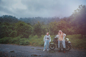 couples of asian biker wearing plastic raincoat stainding beside small enduro motorcycle against rainy cloud on country track thailand