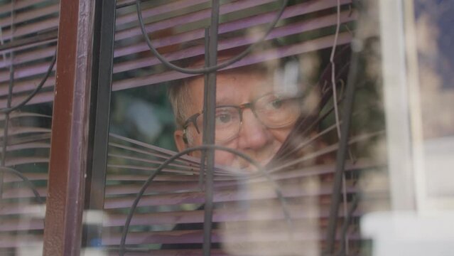 Old man in glasses opens blinds and looks out window, from outside