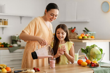 Young woman with her little daughter pouring fresh smoothie into glass in kitchen