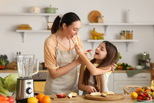 Little Girl With Her Mother Eating Fruits While Making Smoothie In Kitchen