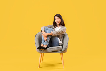 Fototapeta na wymiar Beautiful Asian woman sitting in grey armchair on yellow background