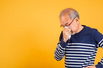 Portrait senior old man blowing nose with tissue studio shot isolated on yellow background, Asian...