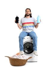 Young man with laundry detergents sitting on washing machine against white background