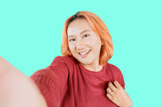 Young Asian woman wearing sweater with red hair standing isolated on tosca background taking selfie photo