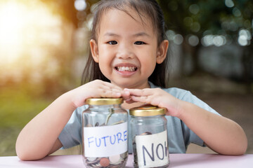 Little Asian girl saving money in Future bottle and now bottle , learning about saving, Kid save money for the future. Money, finances, insurance, and people concept. - 661279229